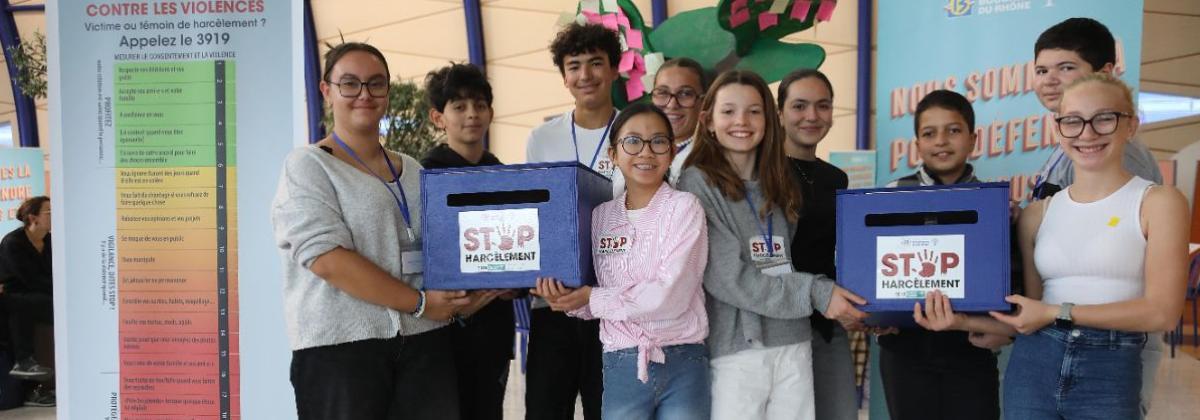 Groupe de sept personnes debout dans un hall lumineux. Elles tiennent deux boîtes bleues avec inscription « STOP Harcèlement ». Derrière, panneaux d’information dont un sur la lutte contre les violences avec une échelle colorée sur le harcèlement et un numéro d’appel.