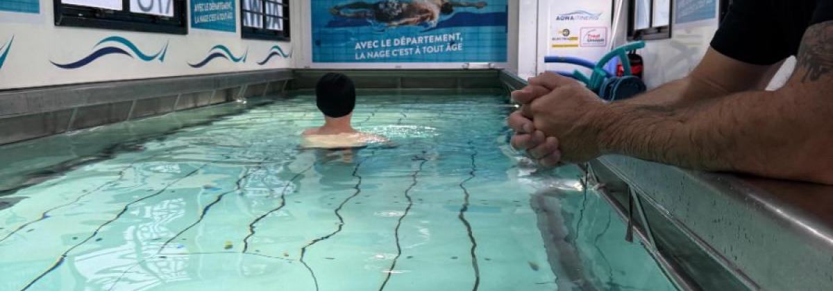Vue rapprochée du bassin intérieur : un adulte appuyé sur le rebord regarde un enfant dans l’eau. Le bassin est long, avec lignes au fond, affiches bleues sur les murs, lumière artificielle, ambiance calme et éducative.
