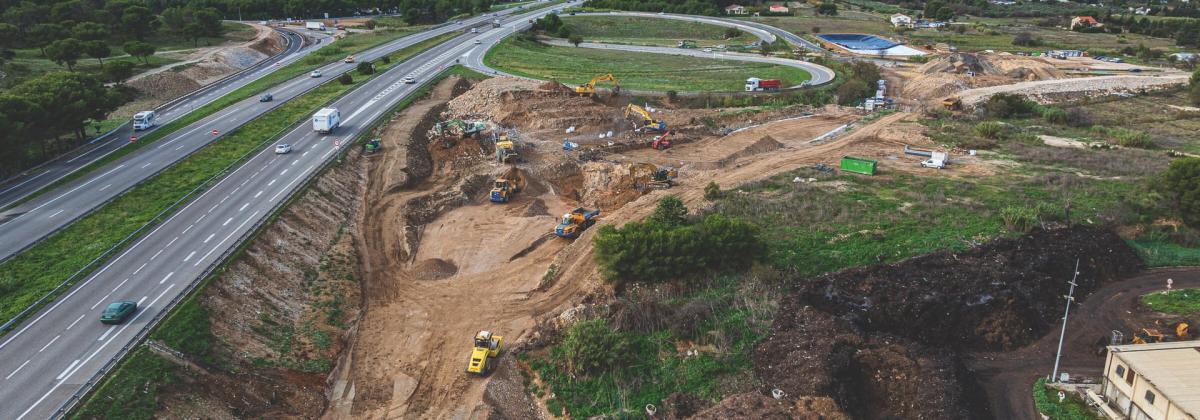 Vue aérienne d’un vaste chantier près d’une autoroute. Aucune personne visible. Plusieurs engins de chantier (pelles, camions, bulldozers) creusent et déplacent la terre. La scène est entourée de collines boisées et de quartiers résidentiels au loin. Le trafic roule sur plusieurs voies adjacentes.
