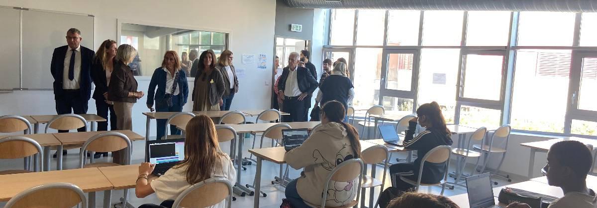 Des élèves sont assis à leur bureau dans une salle de classe, travaillant sur des ordinateurs portables. À l’arrière-plan, un groupe d’adultes observe la scène, probablement en visite pédagogique.