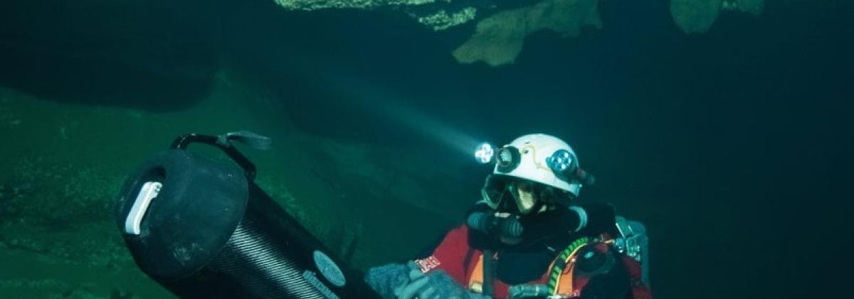 Plongeur sous la roche avec casque et bouteille portant un long appareil en main 