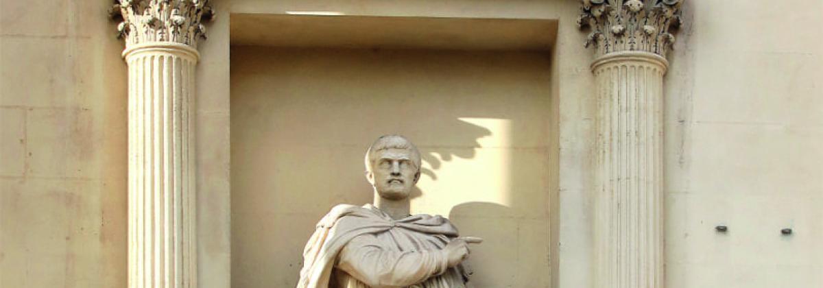 statue d'un homme paré d'une cape par dessus sa tunique et son pantalon, pointant son doigt droit dans une direction tandis que sa main gauche tient un petit trident