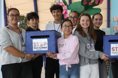 Groupe de sept personnes debout dans un hall lumineux. Elles tiennent deux boîtes bleues avec inscription « STOP Harcèlement ». Derrière, panneaux d’information dont un sur la lutte contre les violences avec une échelle colorée sur le harcèlement et un numéro d’appel.