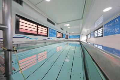Piscine intérieure étroite avec eau claire, murs blancs, fenêtres à cadres noirs et banderoles informatives visibles. Il s'agit de l'intérieur d'un camion piscine. - Agrandir l'image 2 sur 4, fenêtre modale