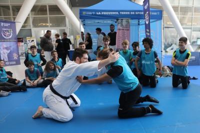 des ados assis sur un tatami en face d'un stand. un adolescent et un adulte se font face à genou, l'un en kimono ceinture noire, l'autre avec un dossard. Ils sont en pleine lutte. - Agrandir l'image 10 sur 15, fenêtre modale