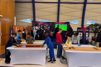 Lors du Mercredi sport et e-sport des Bouches-du-Rhône, enfants et adultes jouent ensemble dans une salle moderne. Autour de tables couvertes de jeux en bois éducatifs, les enfants manipulent, explorent et s’amusent. En fond, deux écrans diffusent des parties de Mario Kart et FIFA, ajoutant une touche numérique à l’ambiance ludique et multiculturelle de l’événement. - Agrandir l'image 1 sur 5, fenêtre modale