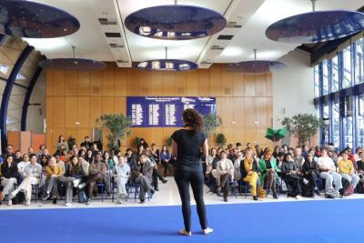 Grande salle lumineuse avec sol bleu et plafond décoré de disques bleus. Une personne debout face à un large public assis sur des chaises fait un discours. Derrière, mur en bois avec panneau bleu listant des noms. Deux grands arbres décoratifs sont visibles. Les participants regardent la personne qui semble présenter ou animer. - Agrandir l'image 2 sur 5, fenêtre modale