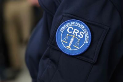 Un adulte en uniforme sombre est montré de près, centré sur la manche. Un écusson rond bleu et blanc y est fixé, portant la mention “OFFICIER DE POLICE JUDICIAIRE – CRS”. L’emblème représente une balance et un flambeau dorés. L’environnement semble être un espace intérieur avec d’autres personnes en tenue professionnelle en arrière‑plan, dans un contexte officiel. Il s'agit de la cérémonie des vœux au personnel des forces de l'ordre et de secours. - Agrandir l'image 15 sur 18, fenêtre modale