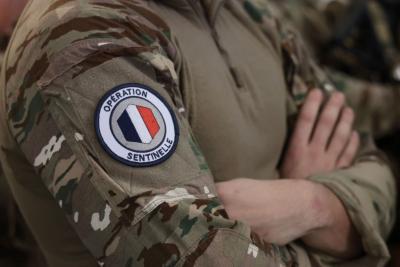 Un adulte en tenue militaire camouflée est photographié de près, les bras croisés. Sur la manche figure un écusson rond indiquant “OPÉRATION SENTINELLE”, autour d’un emblème tricolore bleu‑blanc‑rouge. L’uniforme comporte un motif camouflage complet et un haut beige zippé. L’arrière‑plan montre d’autres personnes en tenue similaire, dans un environnement intérieur. - Agrandir l'image 17 sur 18, fenêtre modale
