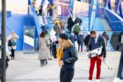 Chanteuse interprétant un titre au micro en extérieur devant un bâtiment bleu, accompagnée d’un musicien jouant d’un keytar parmi les passants - Agrandir l'image 4 sur 20, fenêtre modale