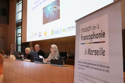 Dans un amphithéâtre institutionnel, trois personnes sont assises derrière une table équipée de micros et d’ordinateurs. Un grand écran projette une présentation. À droite, un panneau annonce « maison de la francophonie à Marseille ». L’ambiance est formelle et organisée. - Agrandir l'image 1 sur 5, fenêtre modale