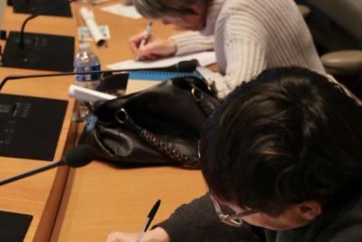 Dans une salle de conférence, plusieurs adultes écrivent concentrés sur des feuilles quadrillées posées devant eux. Ils sont assis côte à côte, penchés sur leur travail. Des micros, carnets et bouteilles d’eau sont disposés sur la longue table en bois. - Agrandir l'image 3 sur 5, fenêtre modale