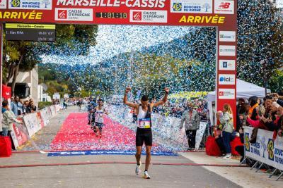 Coureur franchissant la ligne d’arrivée du Marseille-Cassis sous une pluie de confettis bleus, avec un tapis rouge au sol, l’arche d’arrivée et des spectateurs applaudissant en arrière-plan. - Agrandir l'image 10 sur 12, fenêtre modale