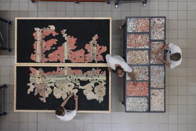 des personnes reconstituent une fresque, morceaux par morceaux sur une table de fond noir. A côté, les morceaux de la fresque ont été triés dans différents bacs, par couleurs. - Agrandir l'image 7 sur 14, fenêtre modale