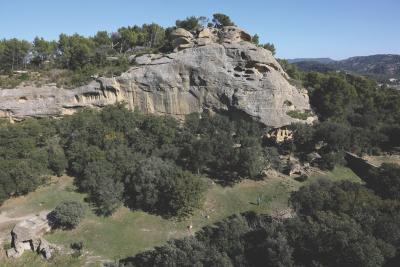 Grande falaise rocheuse beige avec des cavités, entourée de végétation dense et de pins, sous un ciel dégagé. - Agrandir l'image 1 sur 6, fenêtre modale
