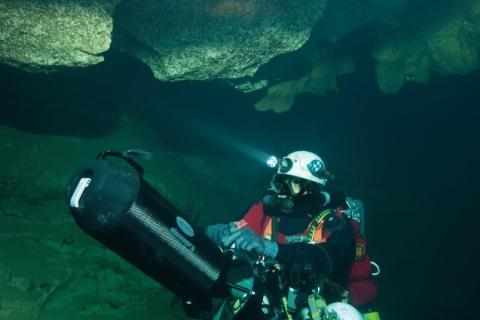 Plongeur sous la roche avec casque et bouteille portant un long appareil en main 