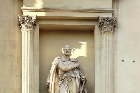 statue d'un homme paré d'une cape par dessus sa tunique et son pantalon, pointant son doigt droit dans une direction tandis que sa main gauche tient un petit trident